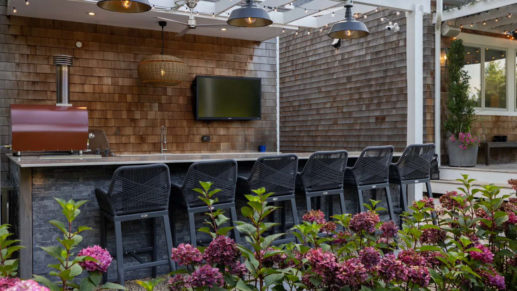 Una barra exterior con asientos oscuros, una pantalla en la pared y plantas con flores.