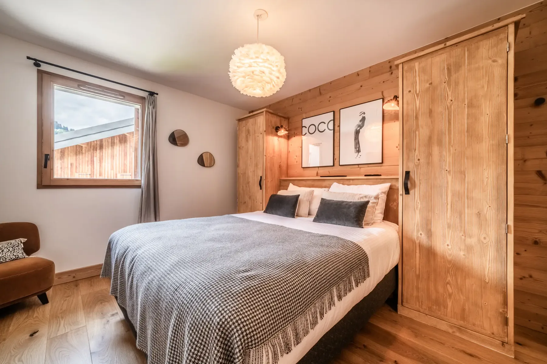 A cozy bedroom with a white bed, gray blanket, wood panel walls, modern artwork, and a fluffy ceiling light.