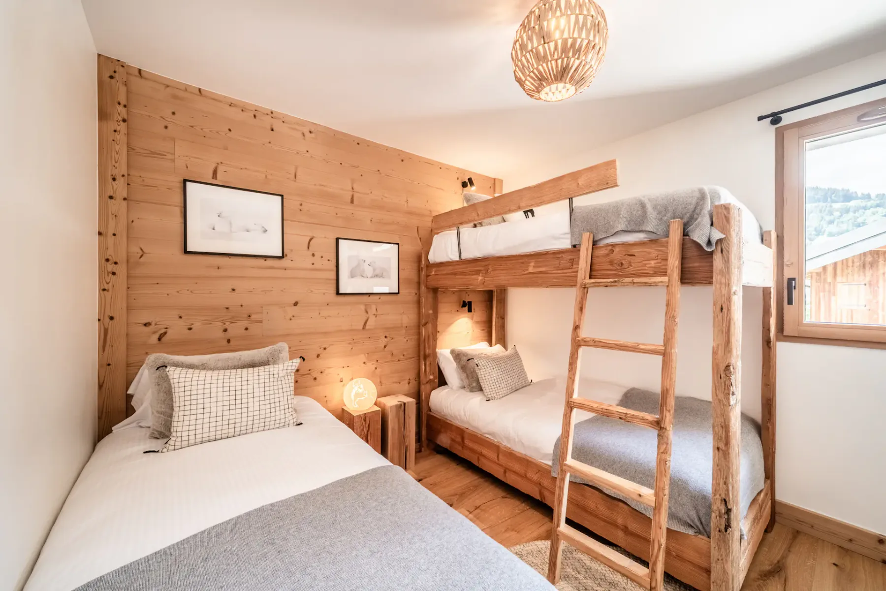 A cozy, well-lit bedroom with wooden walls and furniture, featuring a bunk bed and a single bed, decorated with plush pillows and a teddy bear.