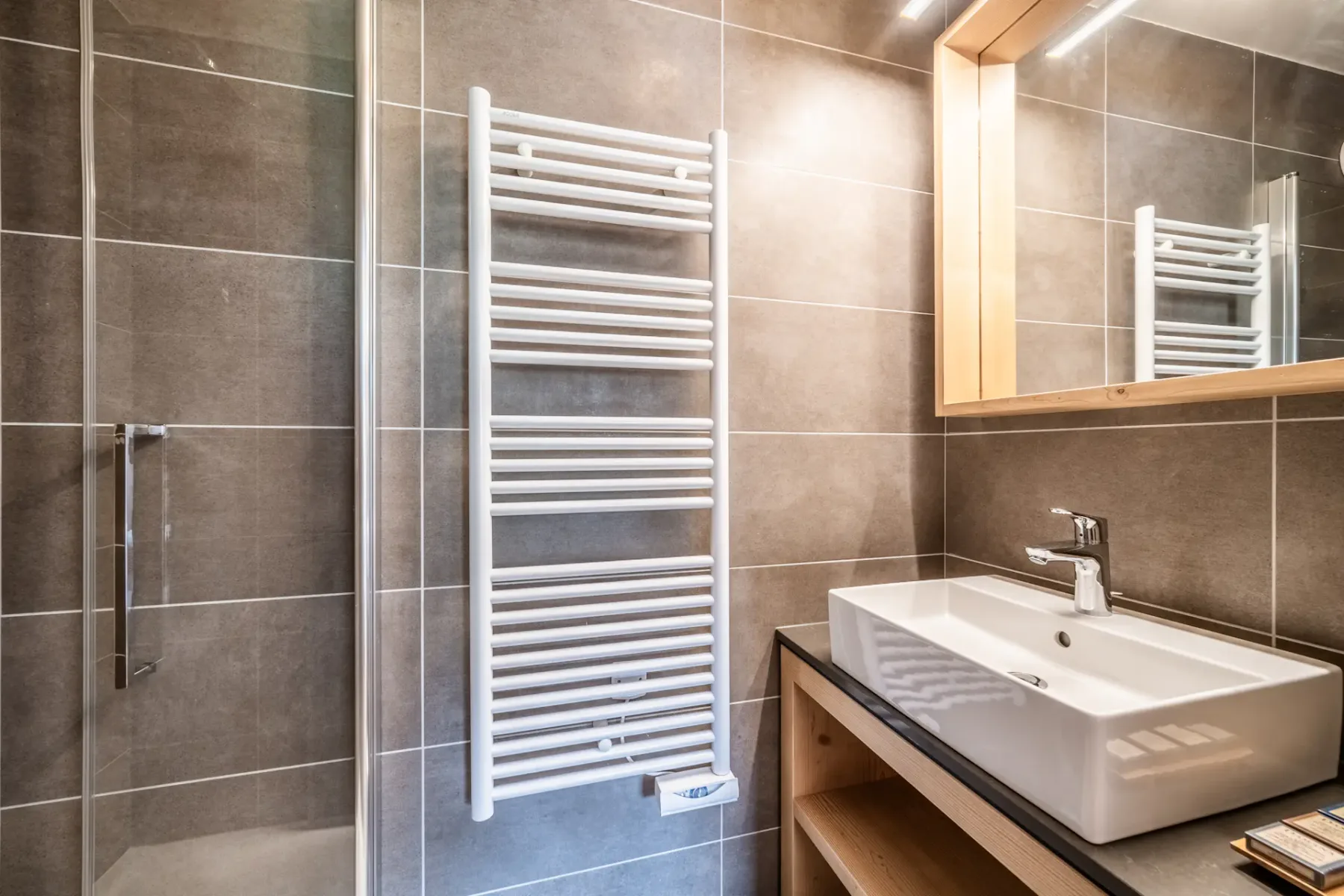 A tiled bathroom with a glass shower, towel radiator, and sink beneath a mirror.