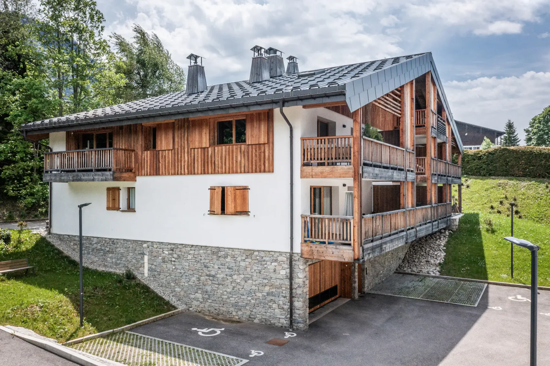A modern chalet with wooden balconies stands beside a paved parking area.