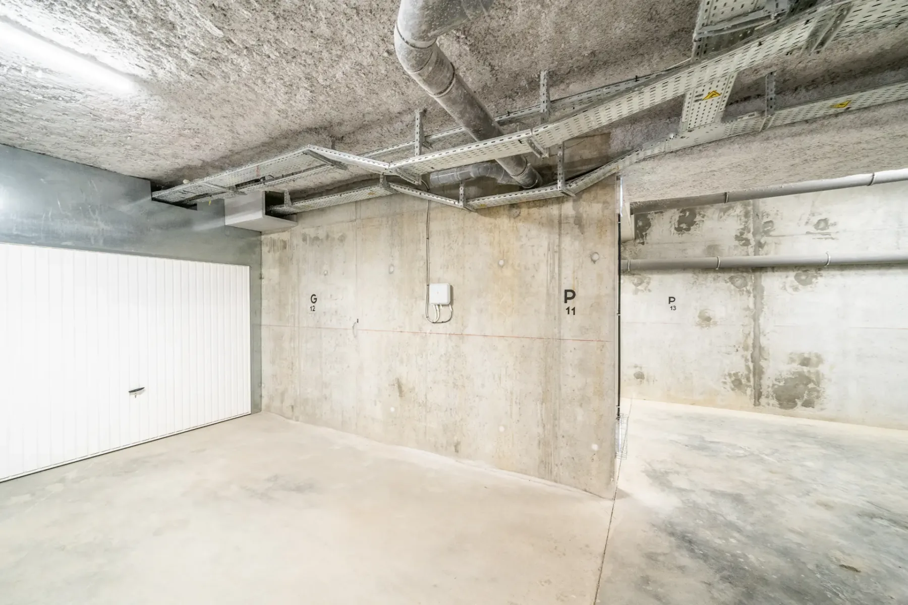 A concrete parking garage with a white sectional door and exposed ceiling pipes.
