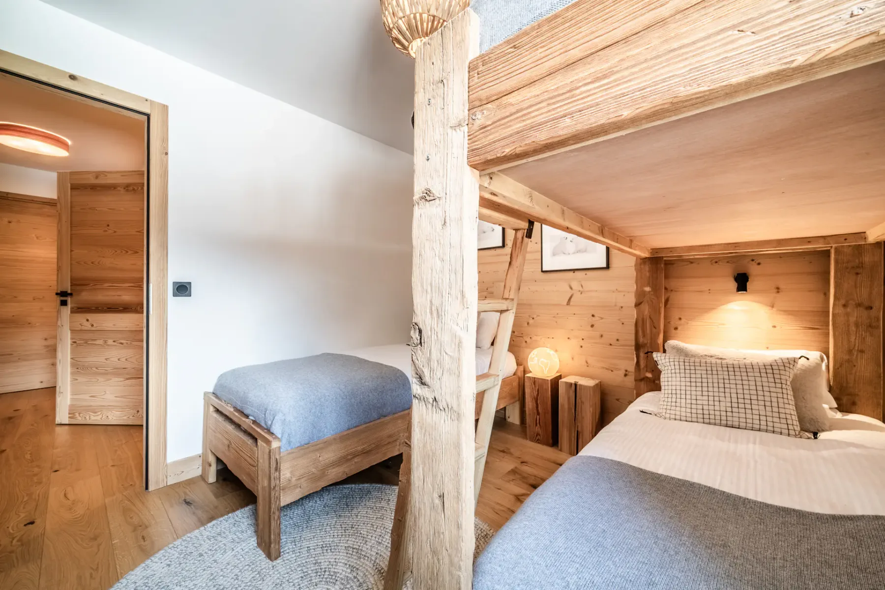 A cozy, well-lit bedroom with wooden walls and furniture, featuring a bunk bed and a single bed, decorated with plush pillows and a teddy bear.