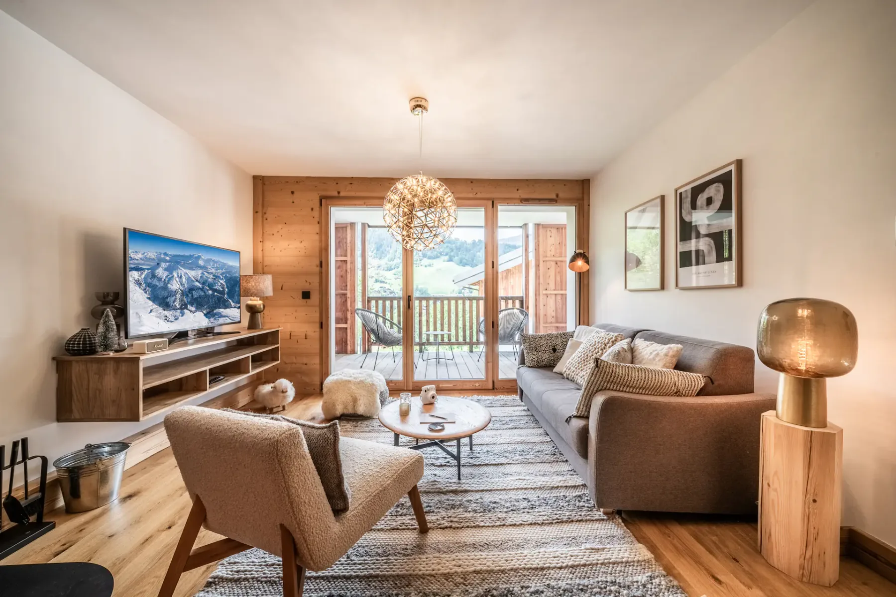 A cozy living room with a modern sofa, wooden flooring, a television, and large doors opening to a balcony with a scenic outdoor view.