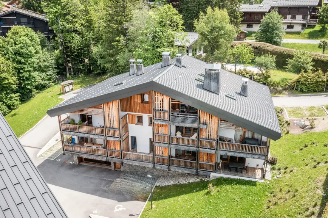 A multi-story chalet building with wooden balconies sits among green trees.