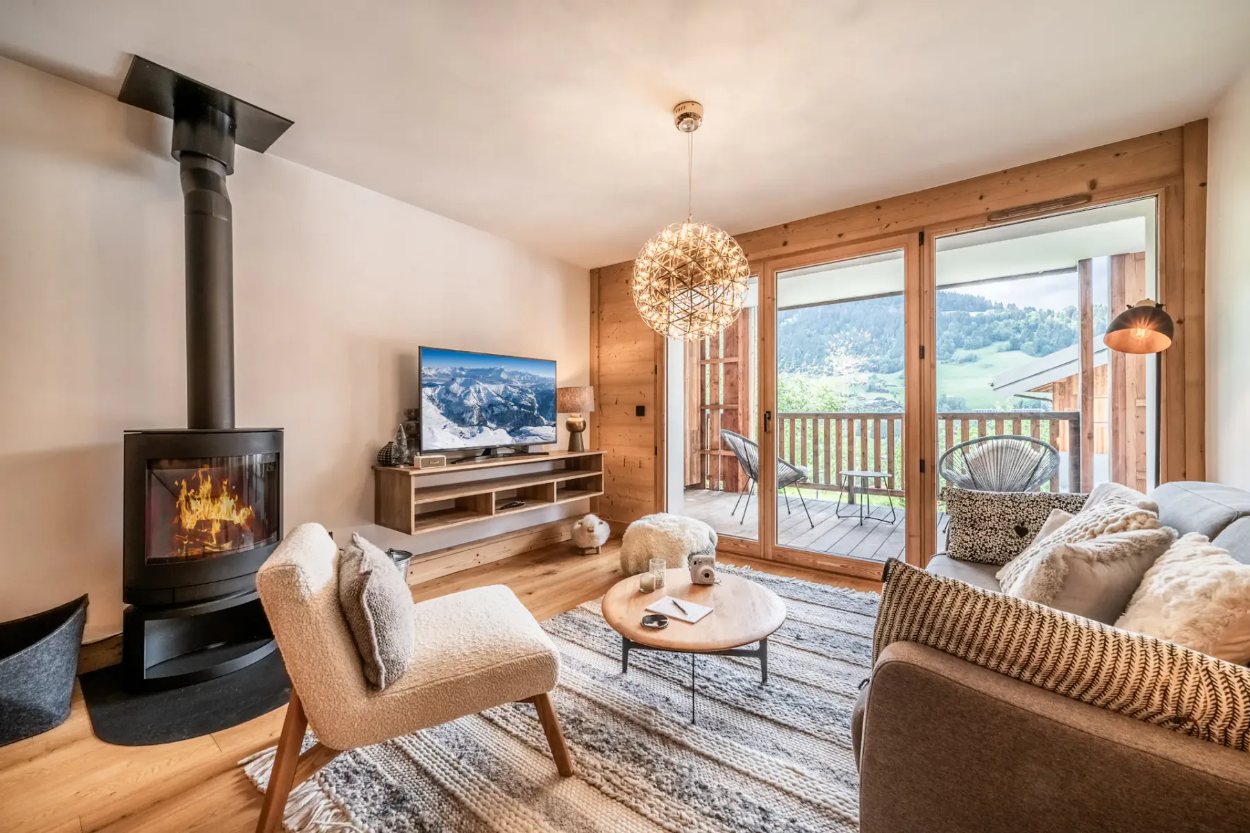 A cozy living room with a modern sofa, wooden flooring, a television, and large doors opening to a balcony with a scenic outdoor view.