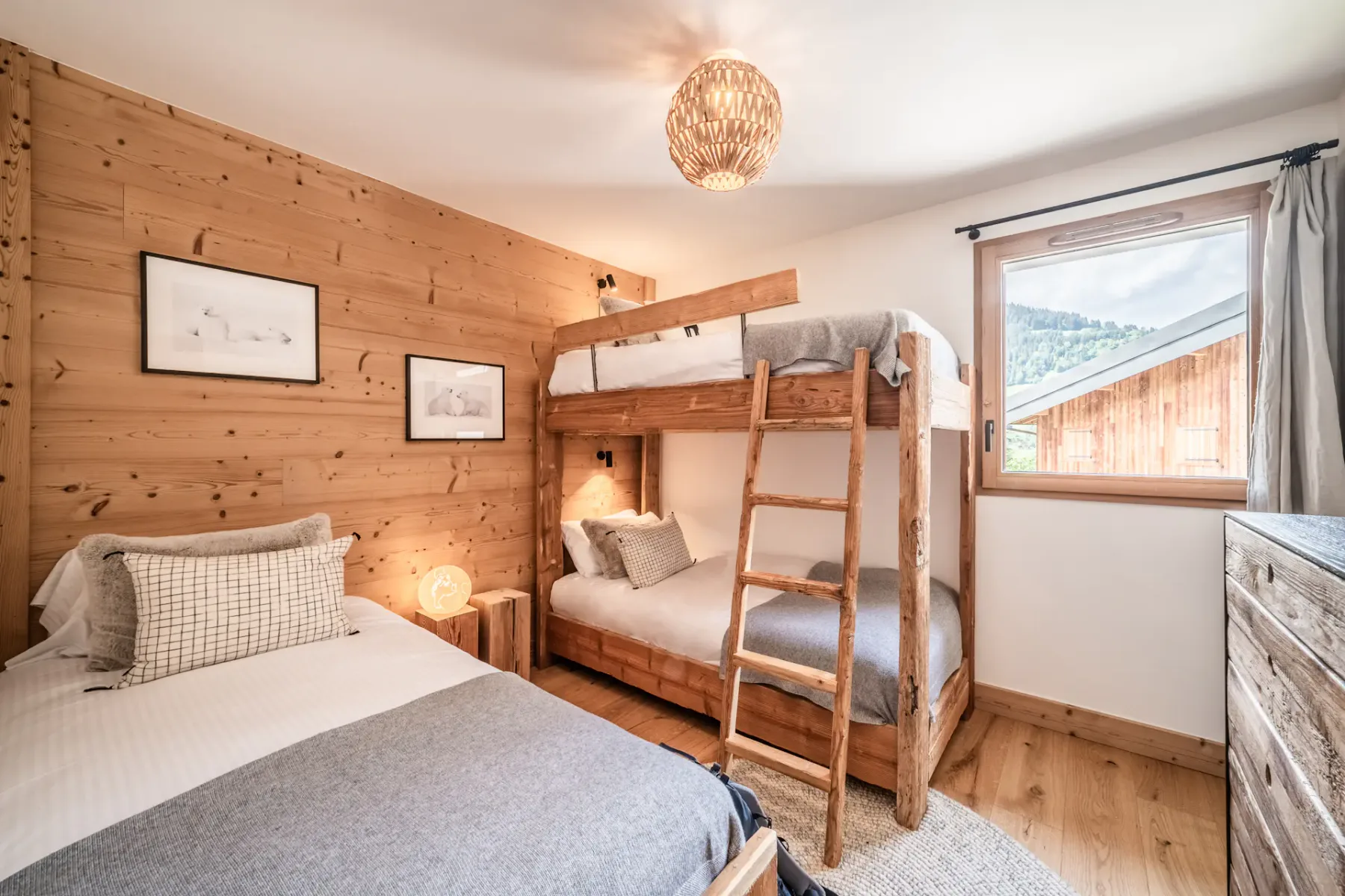 A cozy, well-lit bedroom with wooden walls and furniture, featuring a bunk bed and a single bed, decorated with plush pillows and a teddy bear.