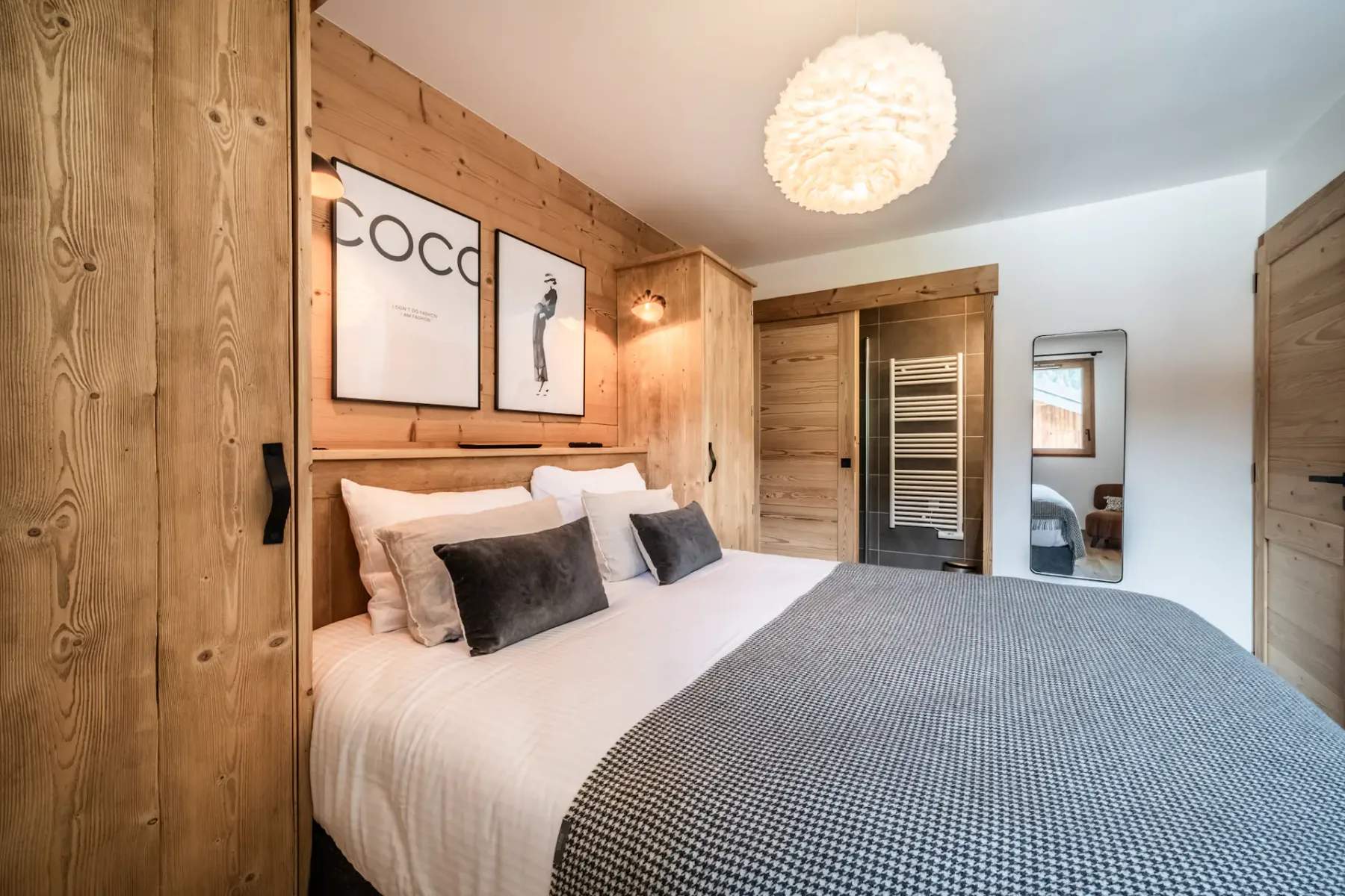 A cozy bedroom with a white bed, gray blanket, wood panel walls, modern artwork, and a fluffy ceiling light.