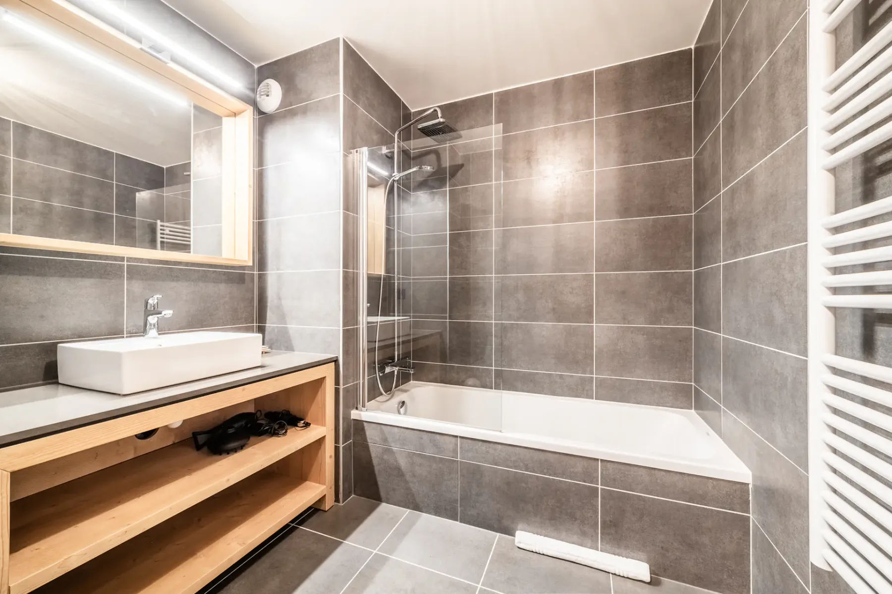 Modern bathroom interior with a bathtub, shower, wooden vanity unit, rectangular mirror, and dark tile walls.