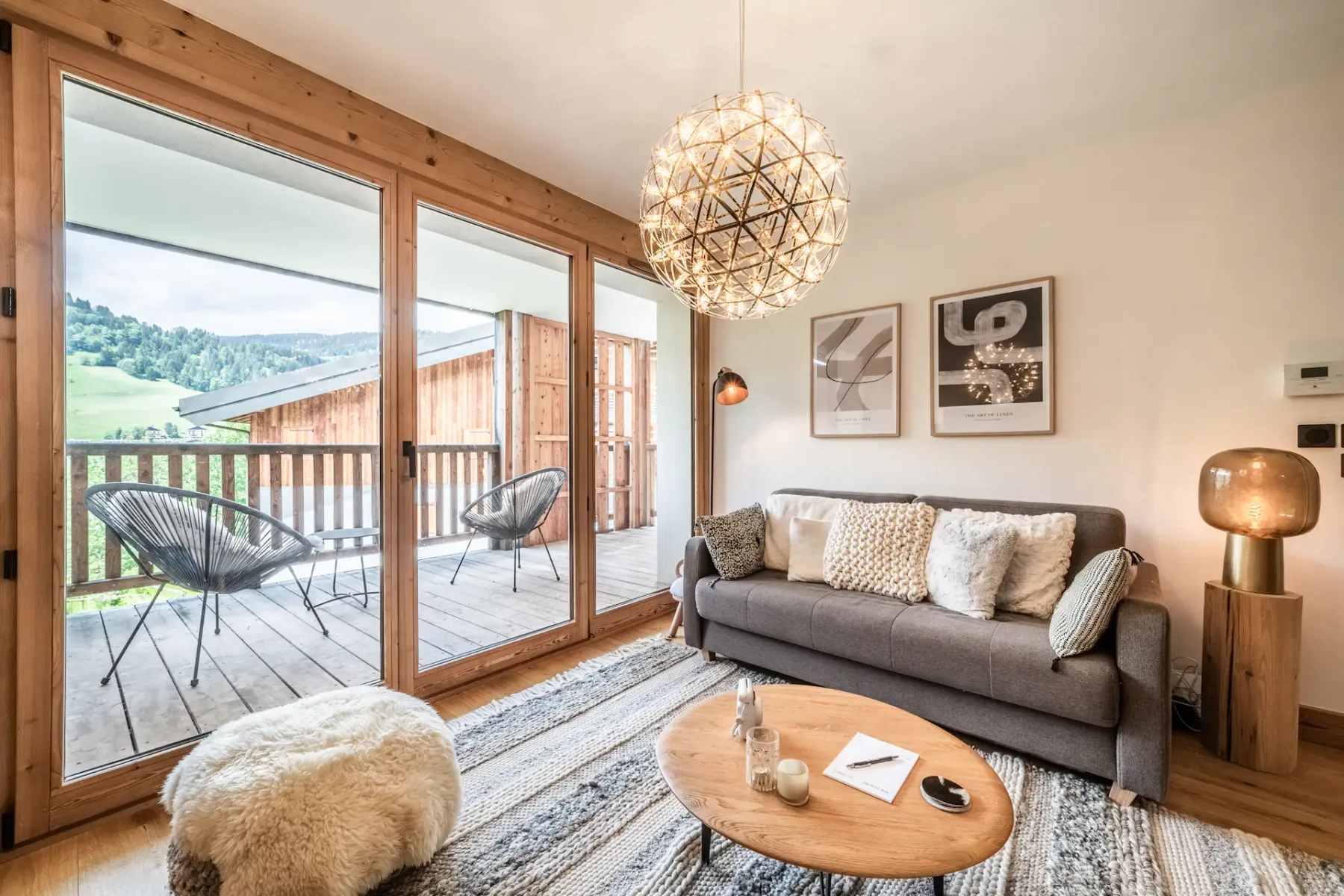 A cozy living room with a modern sofa, wooden flooring, a television, and large doors opening to a balcony with a scenic outdoor view.