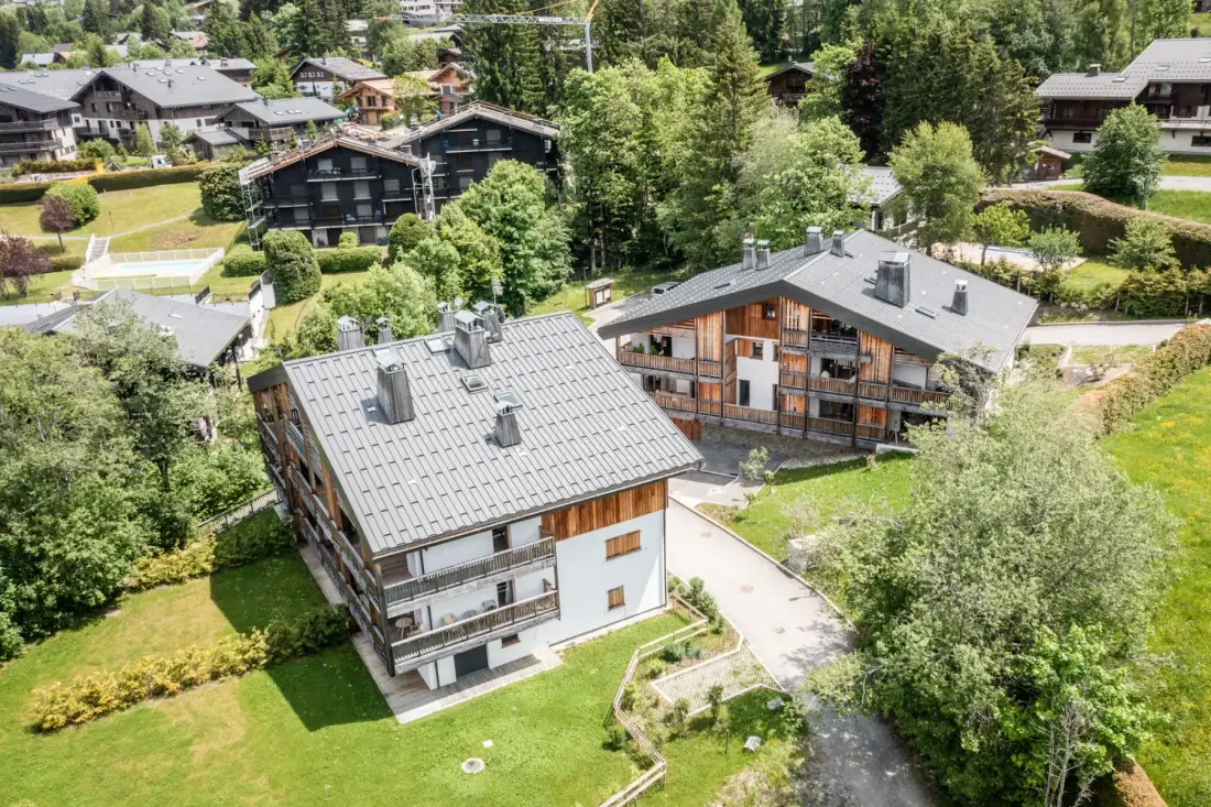 Two chalets sit among green lawns, trees, and other buildings.