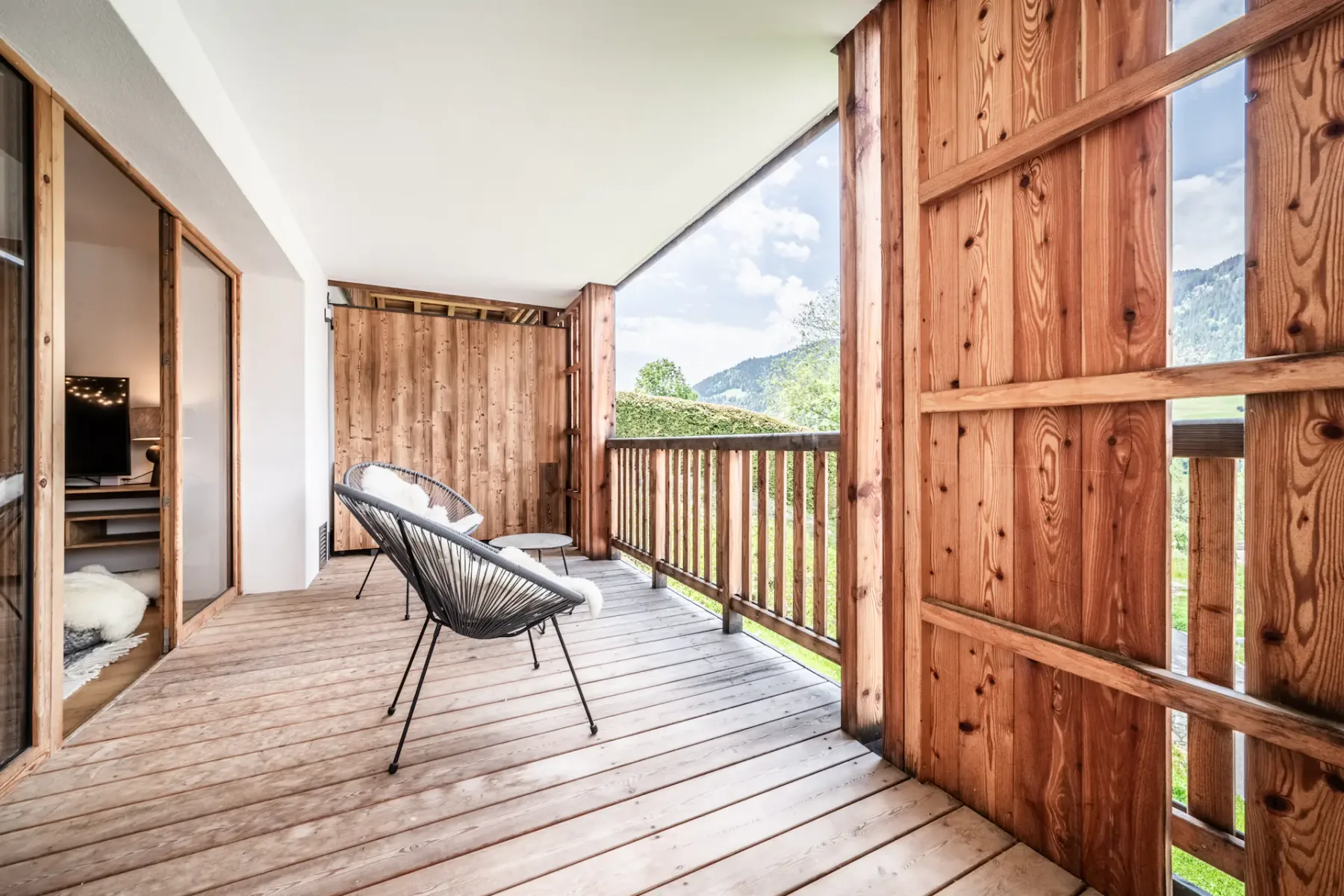 A cozy wooden terrace with a modern chair and table, overlooking green hills, framed by open sliding doors.