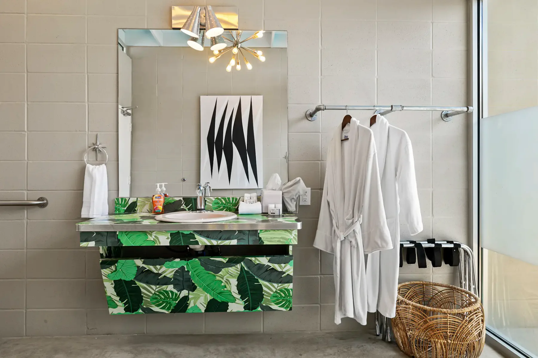 Baño moderno con un mueble de lavabo con estampado de hojas, espejo, una obra de arte y albornoces blancos colgados en un perchero junto a una cesta de mimbre.