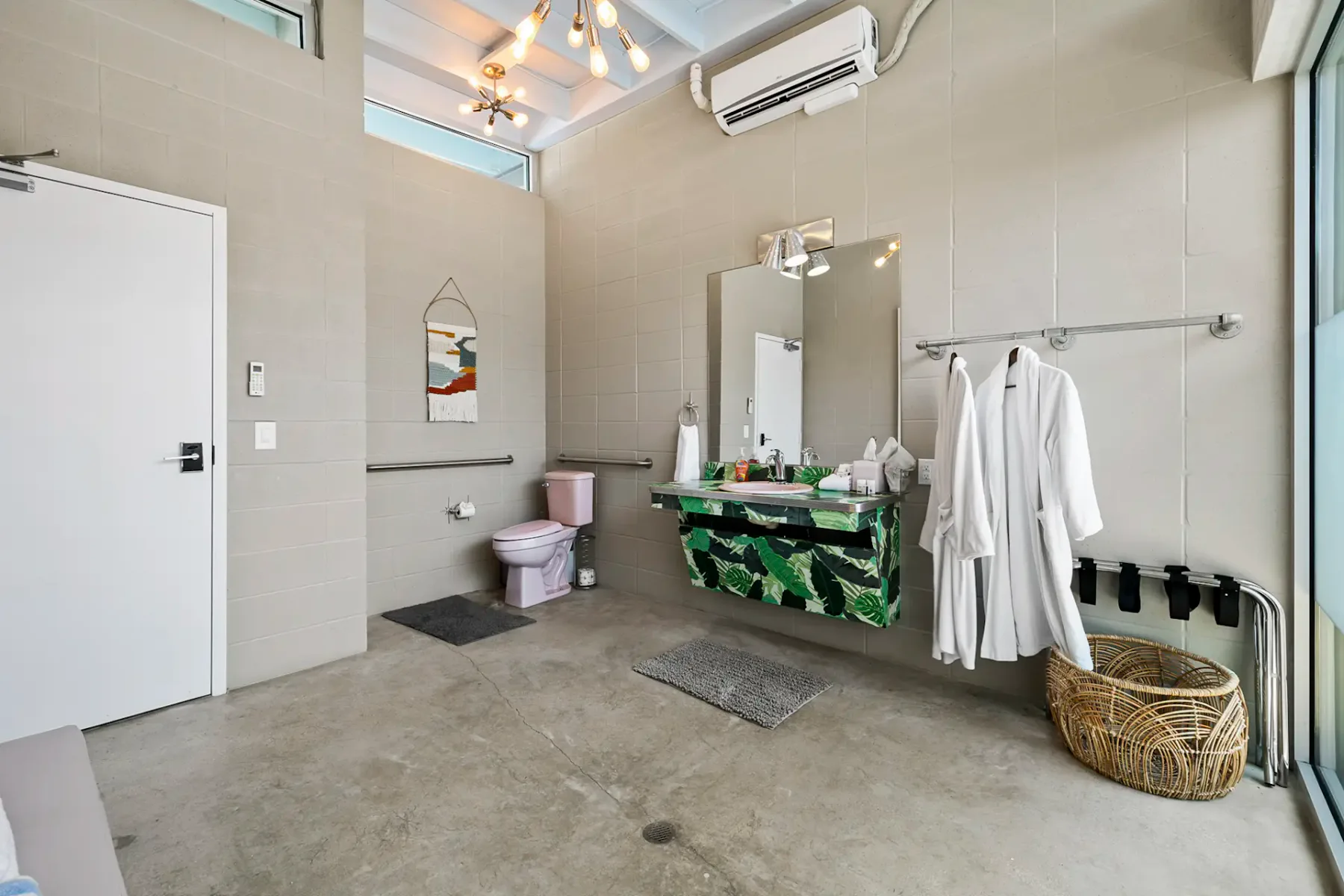 Baño moderno con ducha, inodoro rosa, espejo, dos albornoces blancos y una cesta de mimbre. Paredes de color gris claro y una ventana grande.