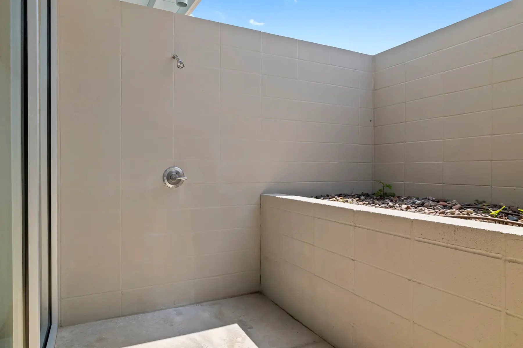 Zona de ducha exterior minimalista con baldosas beige, un cabezal de ducha y una pequeña jardinera con piedras en el lado derecho. Se ve el cielo azul en la parte superior.