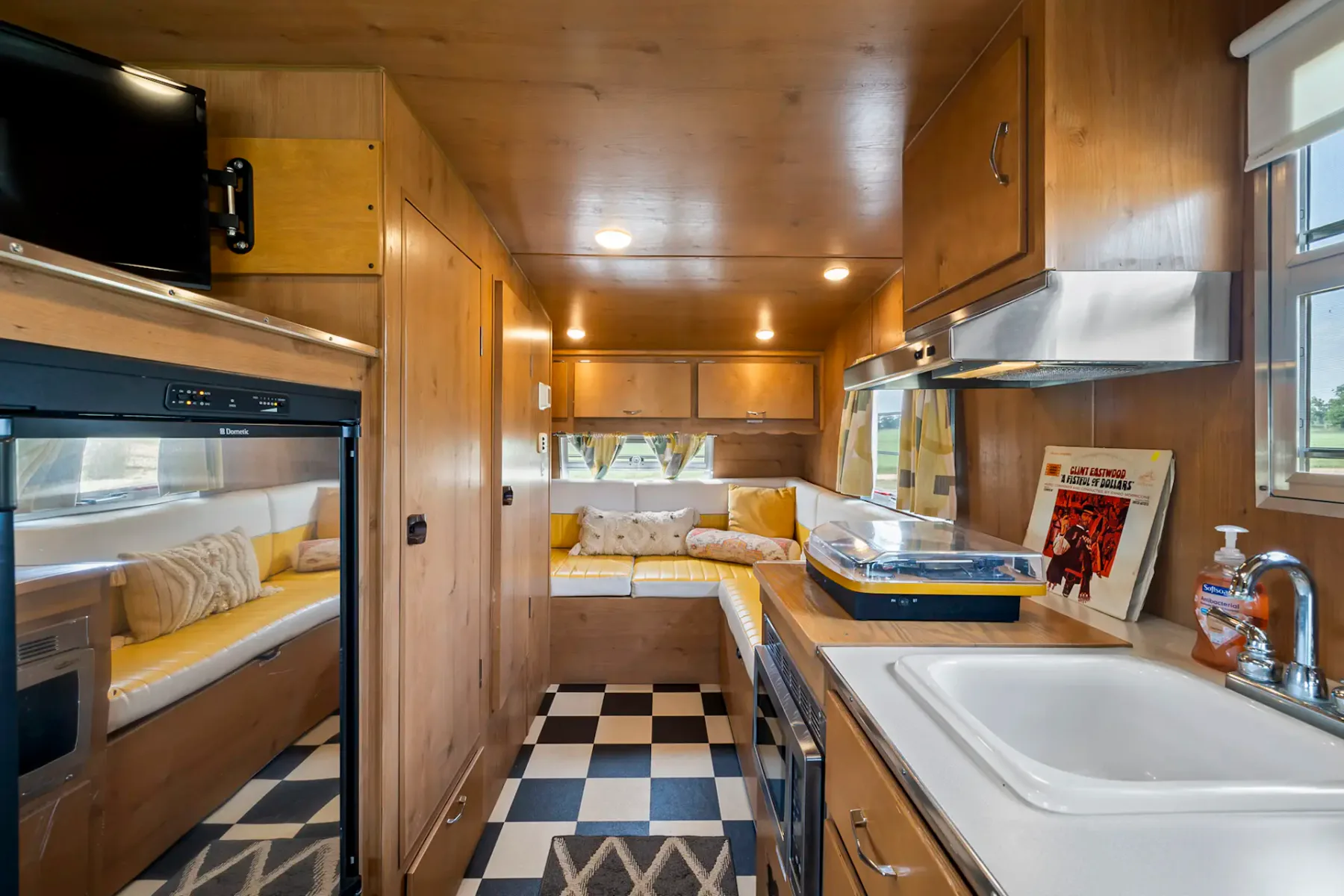 Interior acogedor de una autocaravana con revestimiento de madera, asientos integrados, zona de cocina con fregadero, cocina y frigorífico, suelo de damero y televisión. Luminoso y agradable.