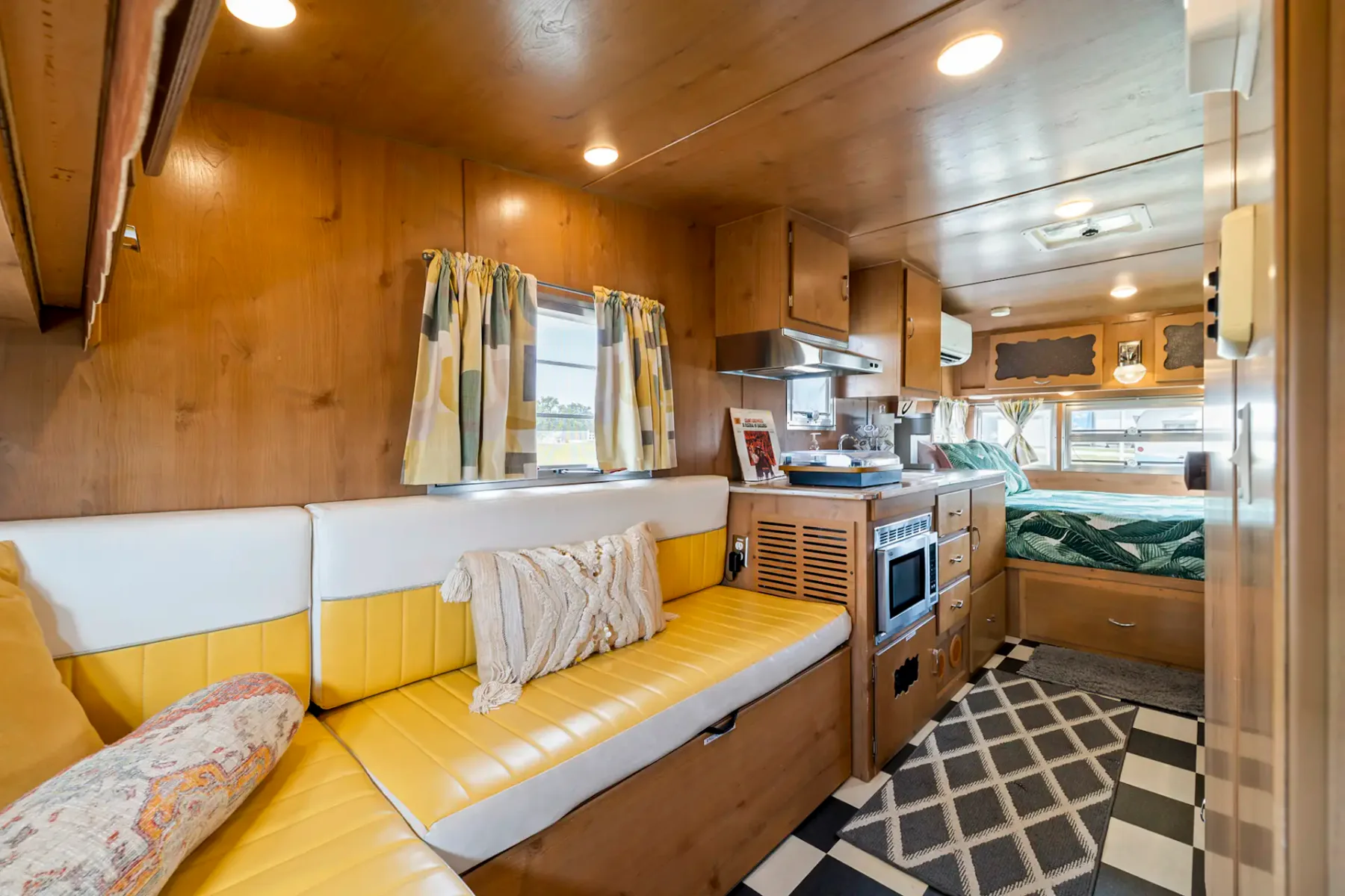 Interior de una caravana acogedora con revestimiento de madera, asientos amarillos, cortinas estampadas, una pequeña cocina y una zona para dormir. Baldosas blancas y negras en el suelo.