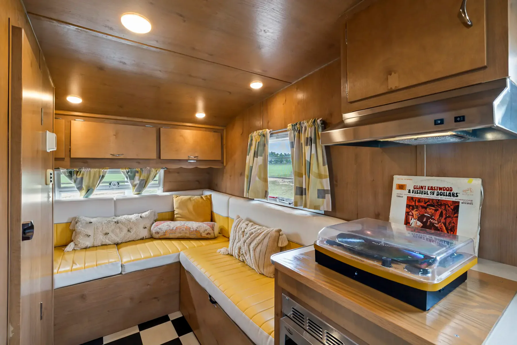 Interior acogedor de una caravana de época con revestimiento de madera, cojines amarillos, un tocadiscos y suelo de damero. Ventana pequeña con cortinas de cuadros.