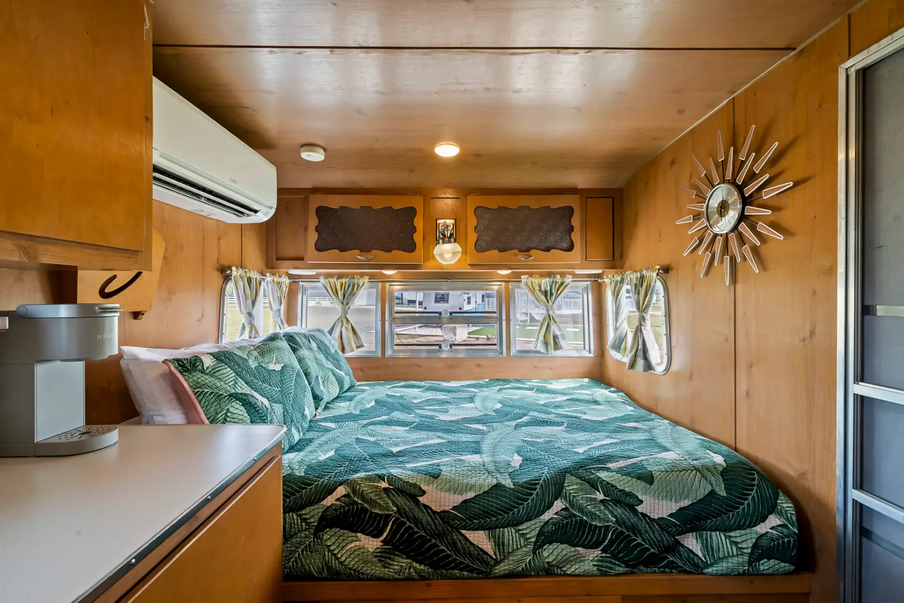 Interior acogedor de una caravana de época con una cama, ropa de cama con motivos tropicales, un reloj en forma de sol y una pequeña cocina con cafetera.