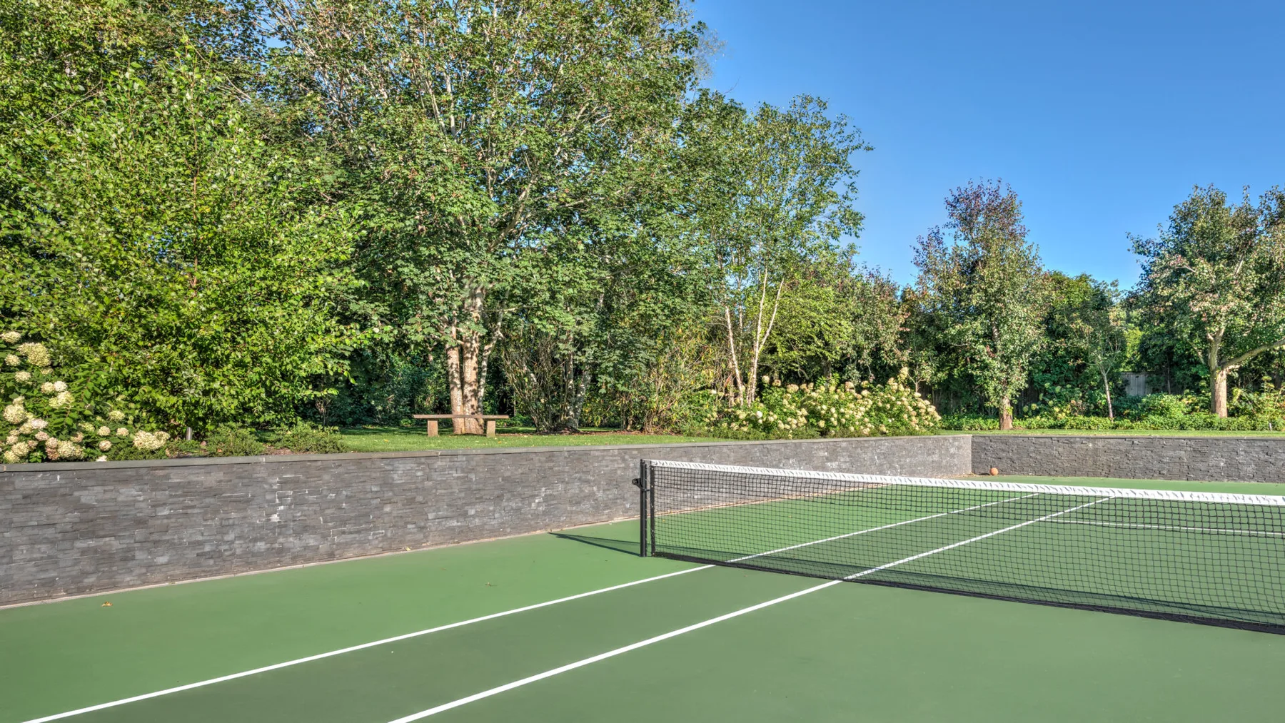 An outdoor tennis court bordered by a low stone wall and trees.