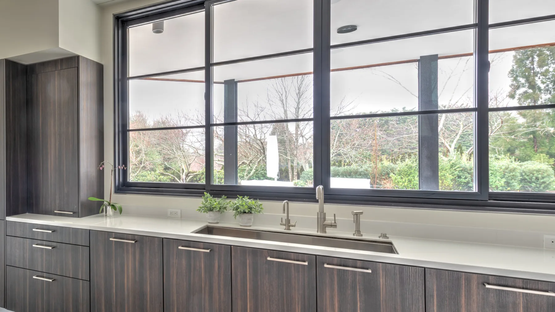A kitchen counter with a sink sits beneath large windows overlooking trees.