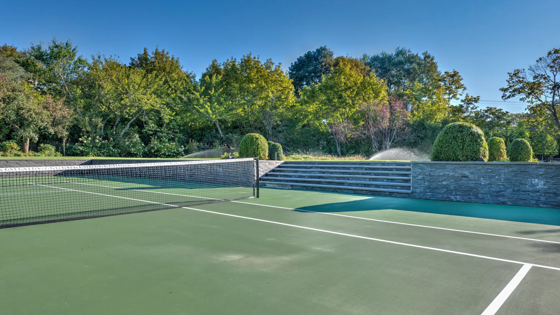 An outdoor tennis court is bordered by trees and a low stone wall.