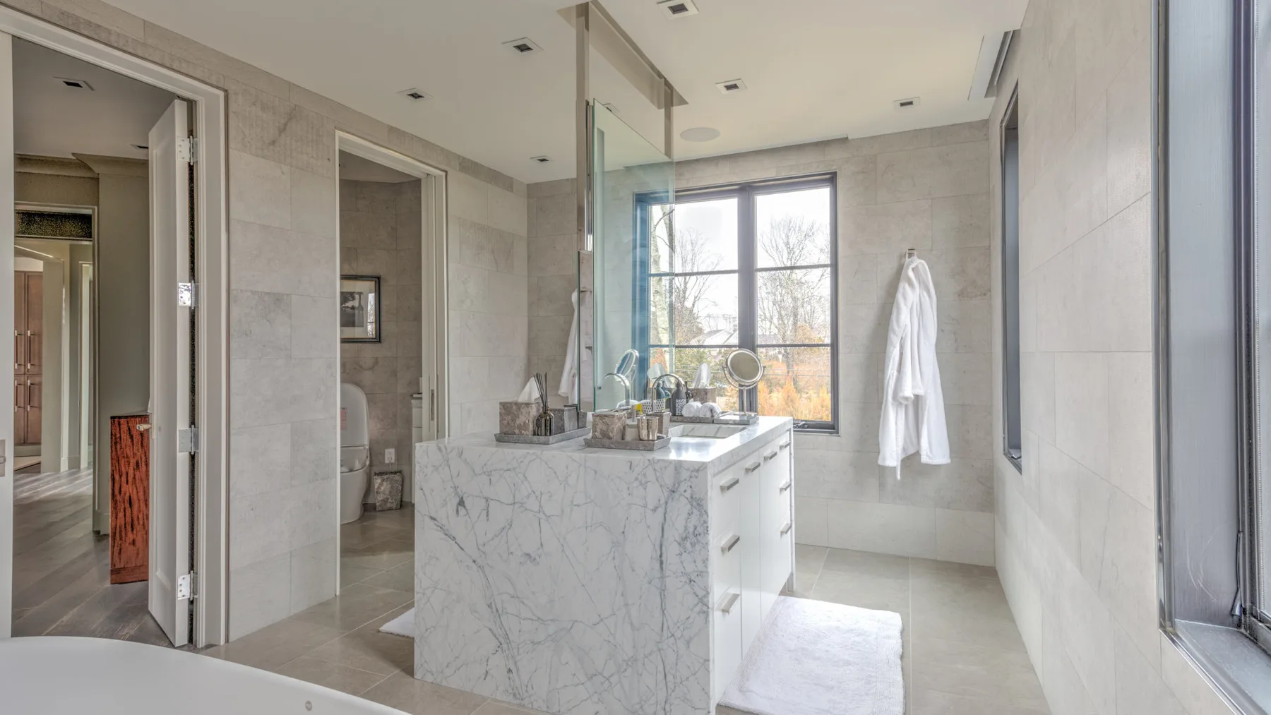 A bright bathroom with a marble vanity, large window, and hanging robe.
