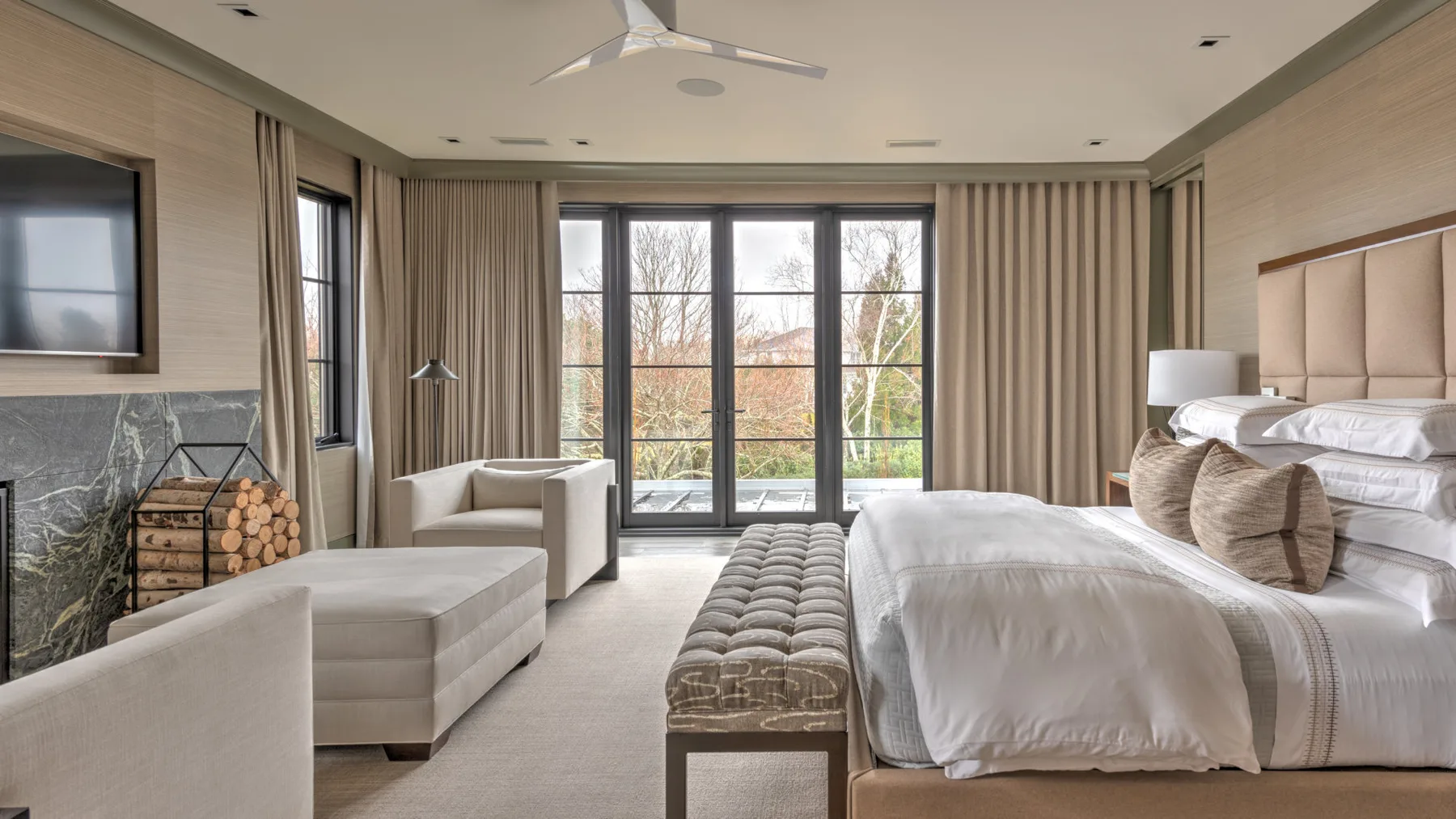 A bedroom with a large bed, a tufted bench, and seating beside tall windows.