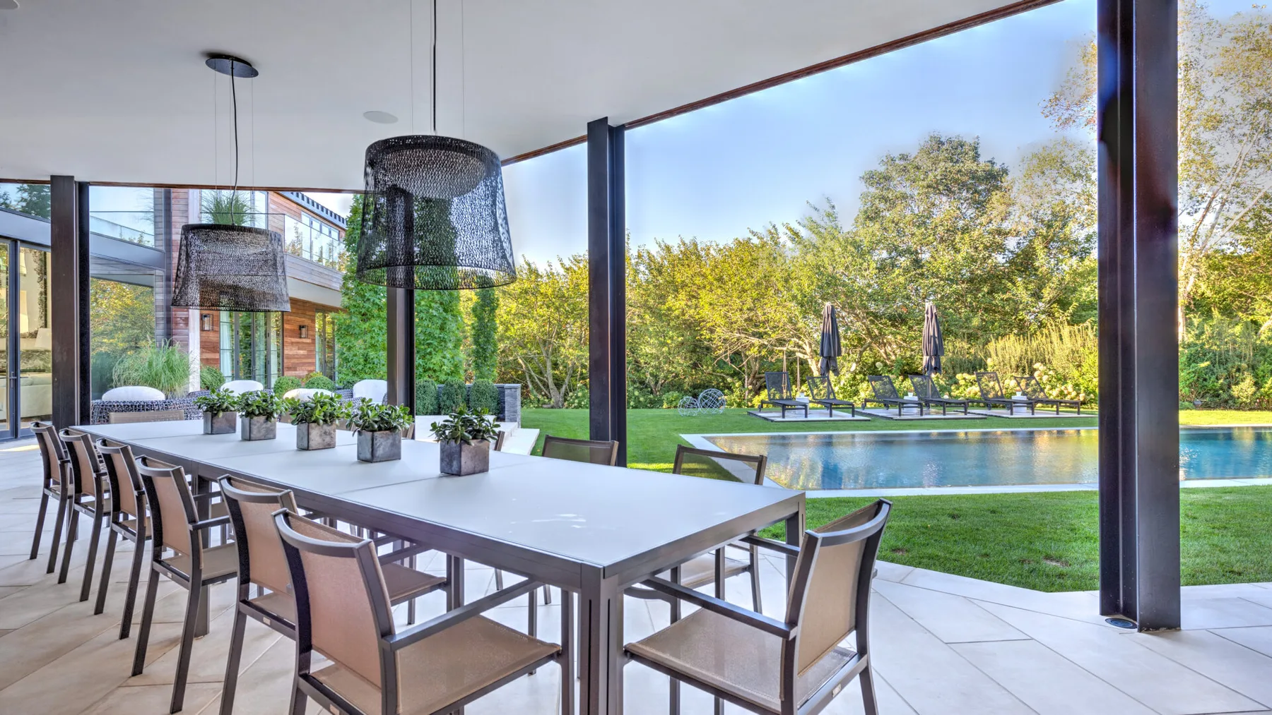 A covered outdoor dining area overlooks a lawn, trees, and a swimming pool.