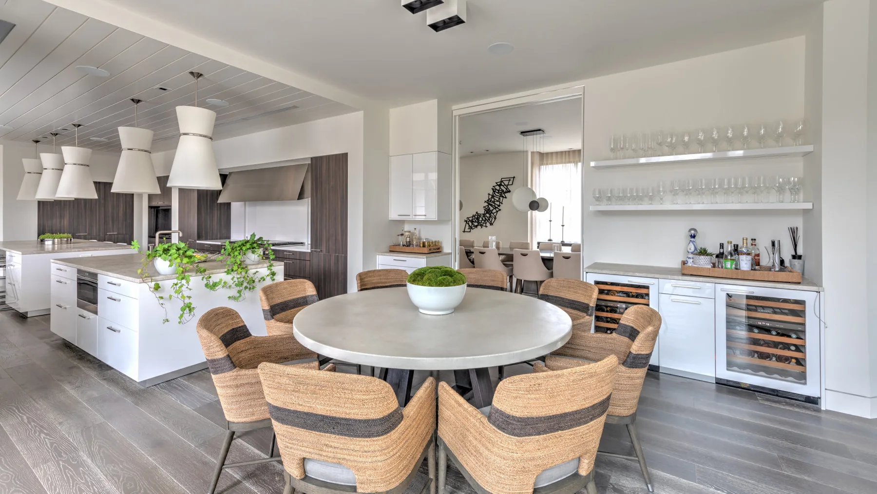 A bright dining area has a round table with woven chairs beside an open kitchen.