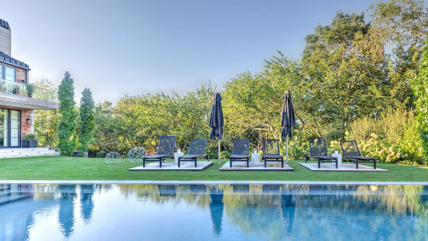 An outdoor swimming pool borders a lawn with lounge seating and trees.