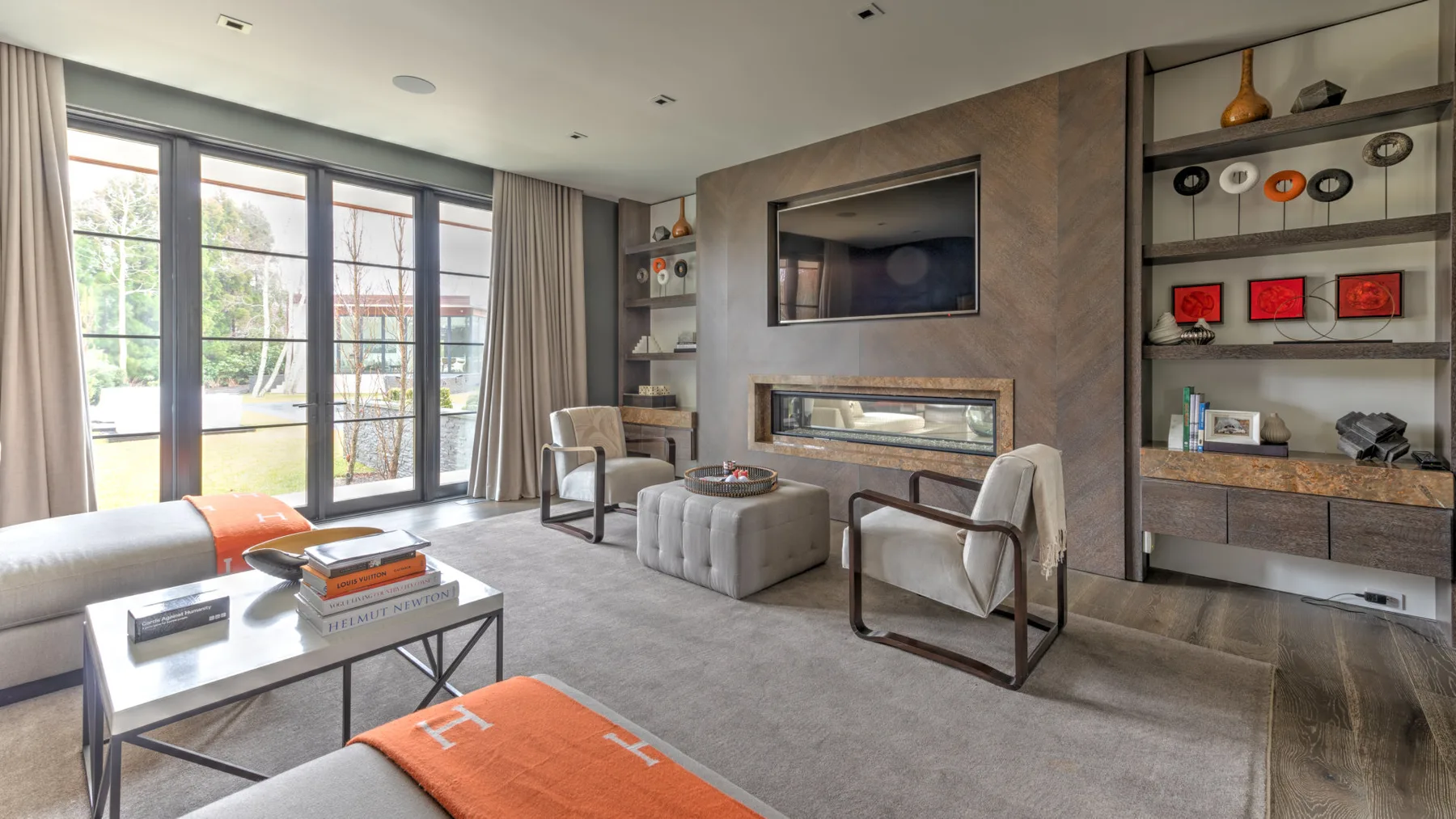A living room with a television above a fireplace and seating by large windows.