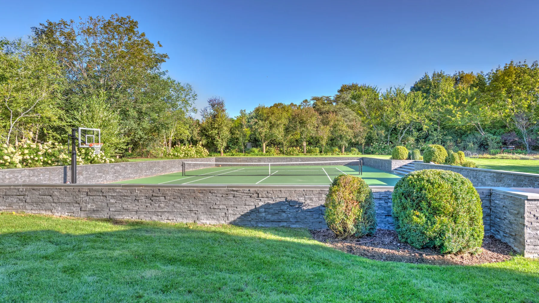 A landscaped outdoor tennis court sits among trees and shrubs.