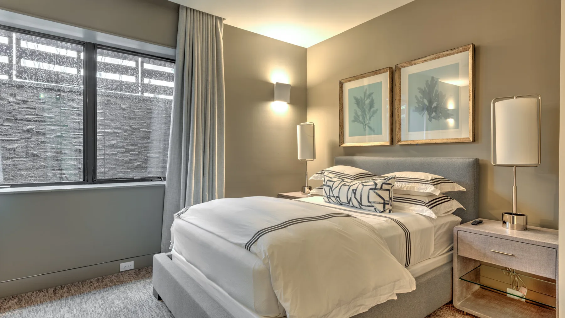 A bedroom with a neatly made bed, framed botanical artwork, and bedside lighting.
