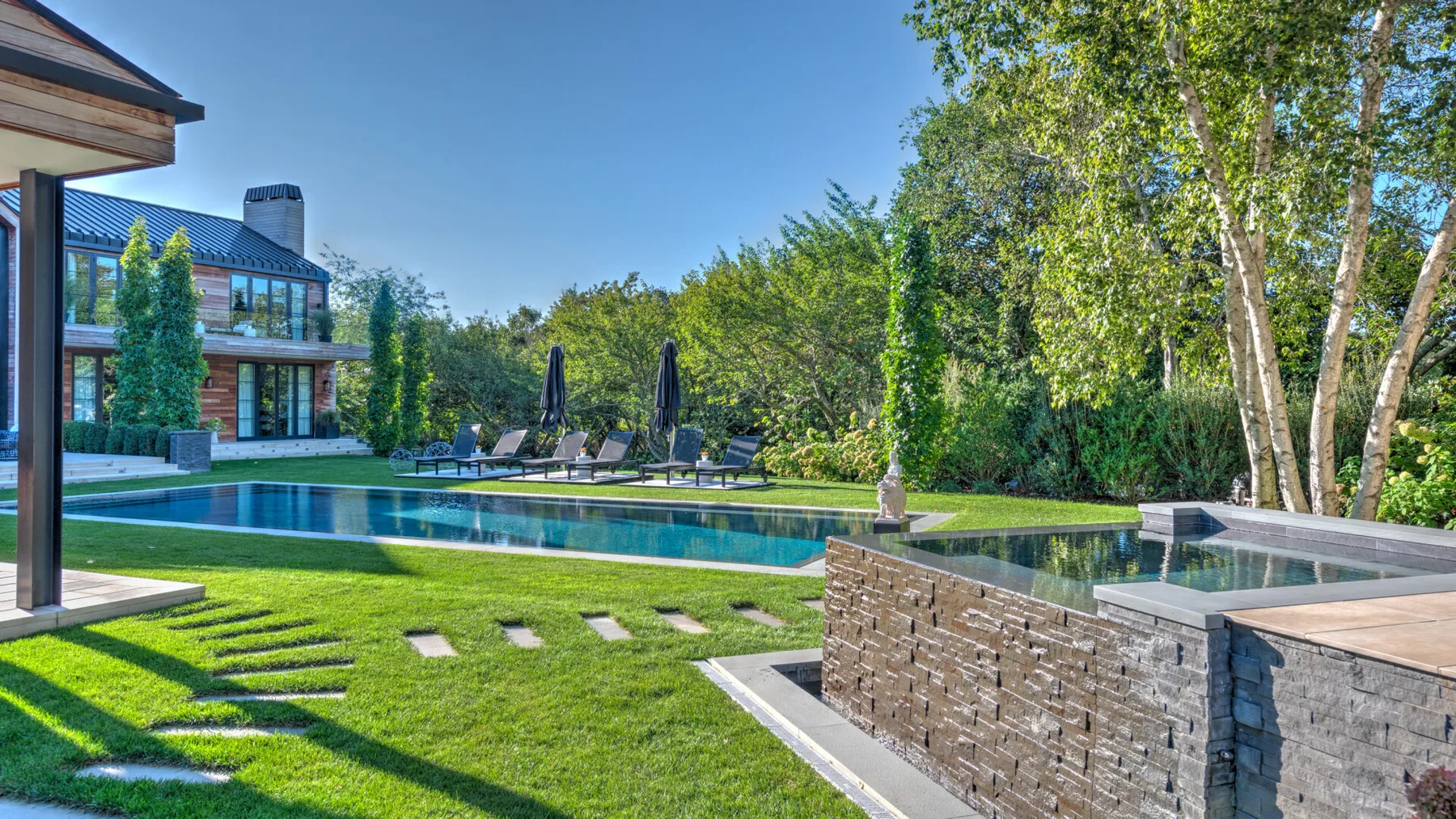 A landscaped outdoor pool beside a modern house, with lounge seating and mature trees.