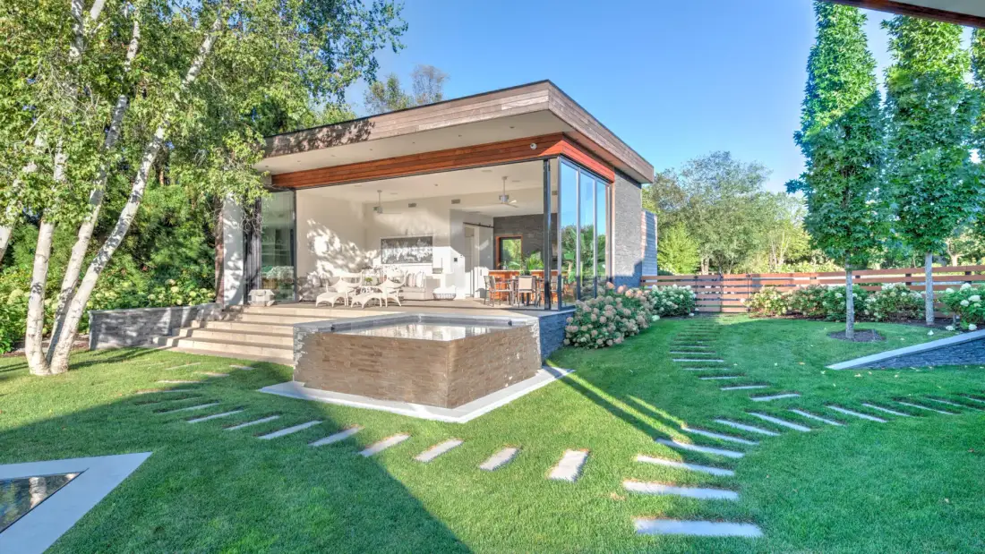 A modern glass-walled pavilion opens onto a lawn with stepping stones.