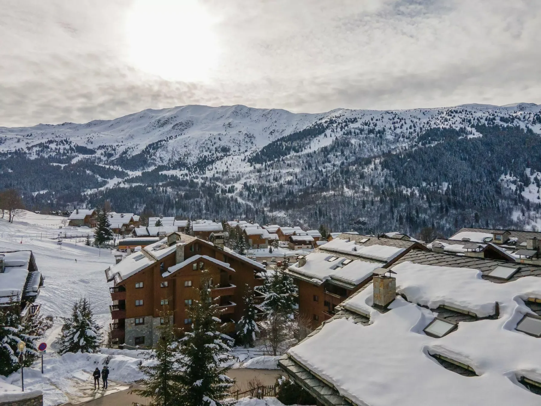 Snow-covered alpine buildings sit below a forested mountain range beneath a cloudy sky.