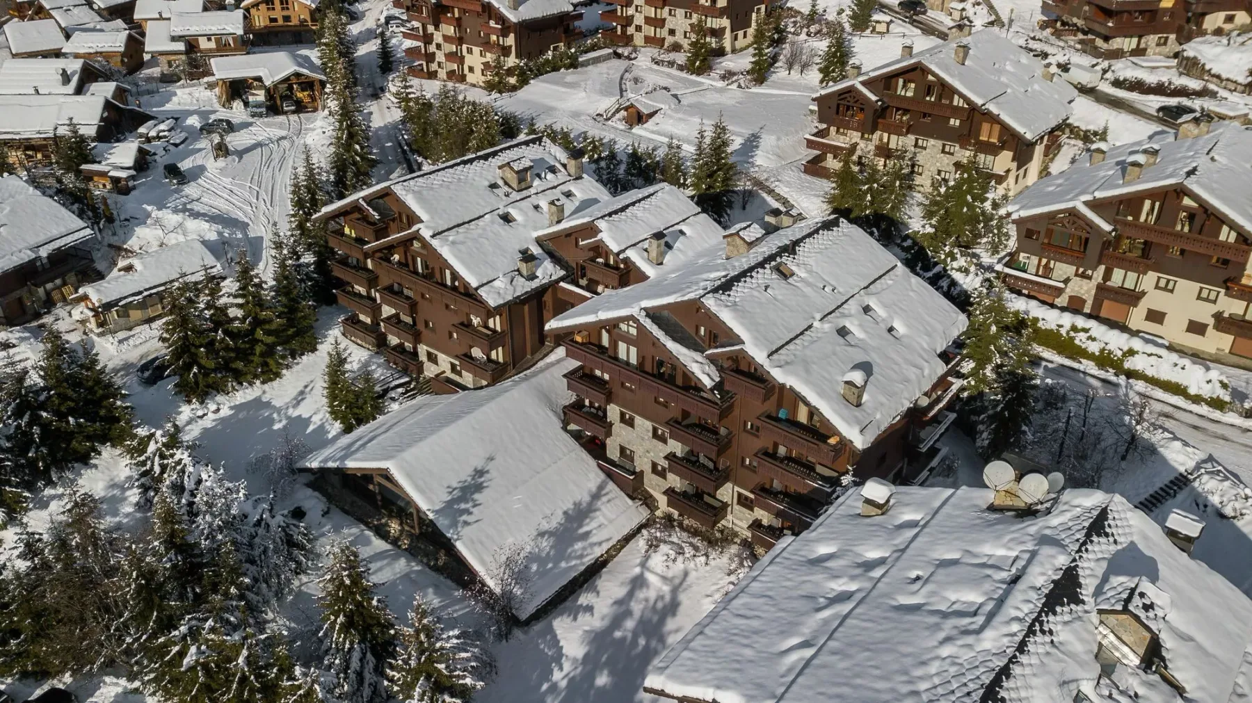 Snow-covered chalet-style buildings sit among evergreen trees in a mountain village.