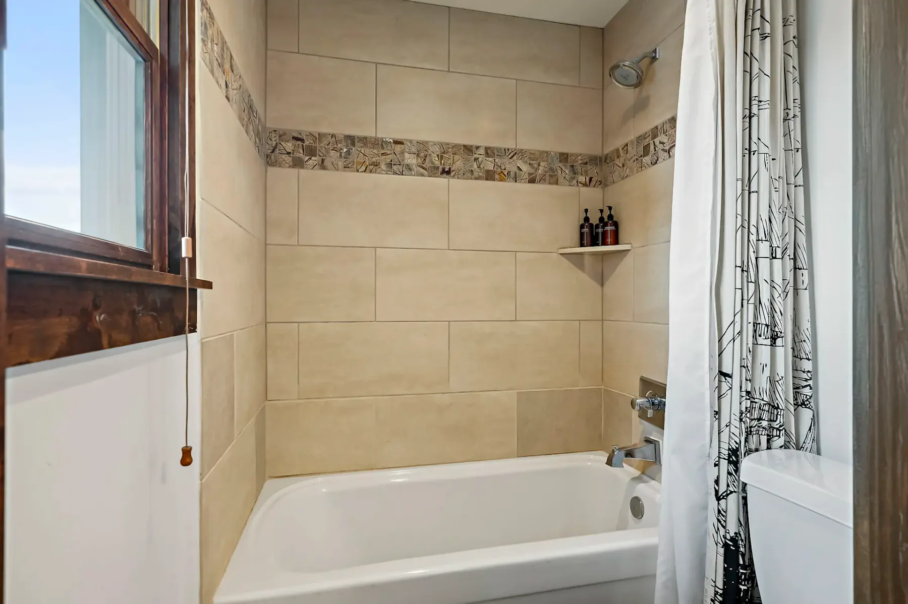 Baño con bañera blanca, paredes revestidas de azulejos beige, cabezal de ducha y cortina de ducha estampada. Hay una ventana a la izquierda.
