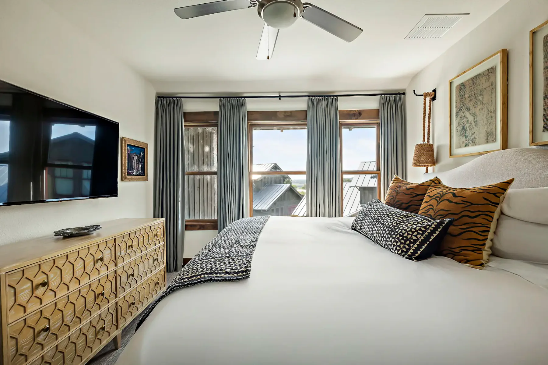 Dormitorio moderno con una cama grande, almohadas estampadas, un televisor montado en la pared, una cómoda de madera, un ventilador de techo y grandes ventanas con cortinas grises.