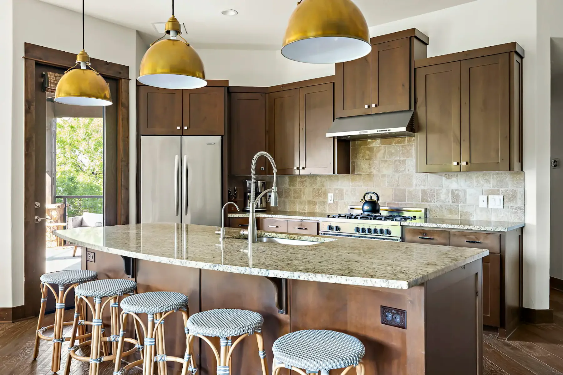 A modern kitchen with wooden cabinets, marble countertops, and three gold pendant lights above a curved island with four cushioned stools.