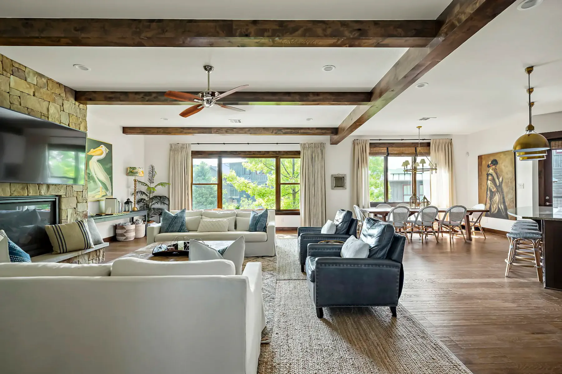 Spacious living room with wooden beams, ceiling fan, cozy seating, stone fireplace, large windows, dining area, and decorative lighting.