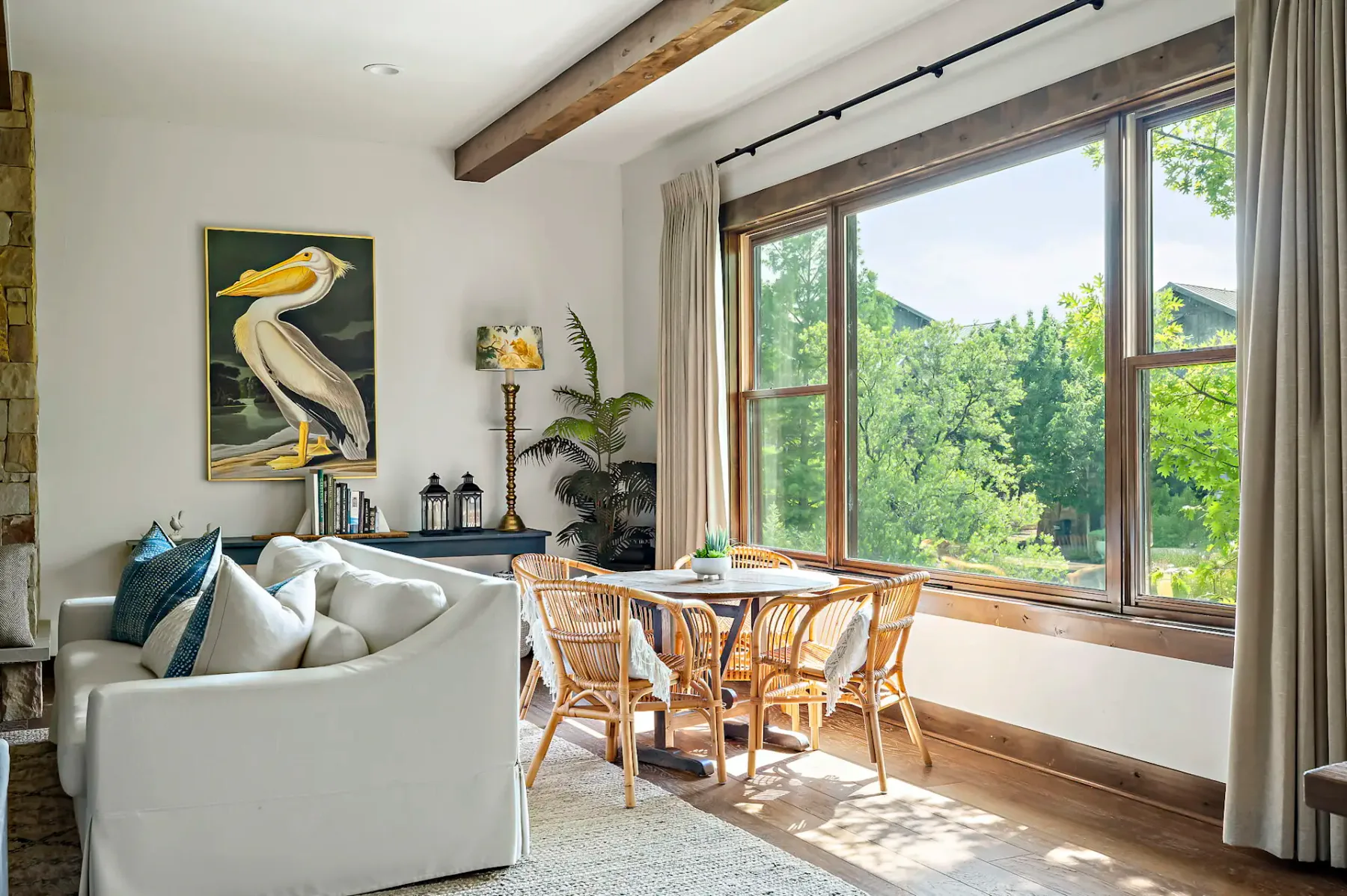 Sala de estar luminosa con una gran ventana, sillas de mimbre alrededor de una mesa, un sofá blanco y una obra de arte con pelícanos en la pared. En el exterior se aprecia una exuberante vegetación.