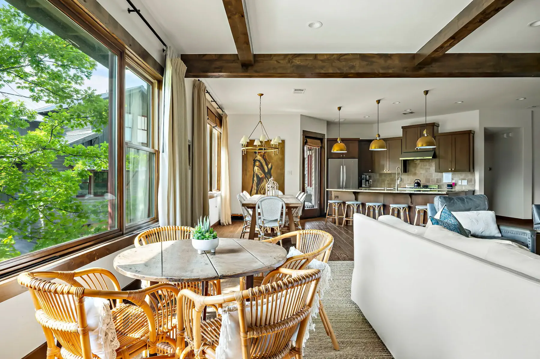 Open living area with wicker chairs, a round table, sectional sofa, and a modern kitchen. Large window offers a view of lush greenery.
