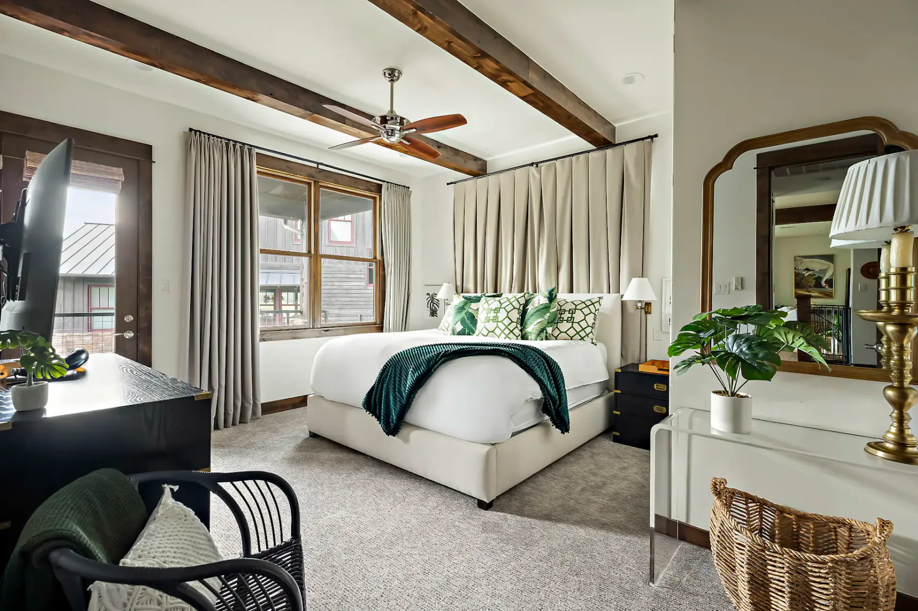 Dormitorio acogedor con una cama con almohadas verdes y una manta, vigas de madera en el techo, una ventana con cortinas y una planta en maceta sobre un escritorio.