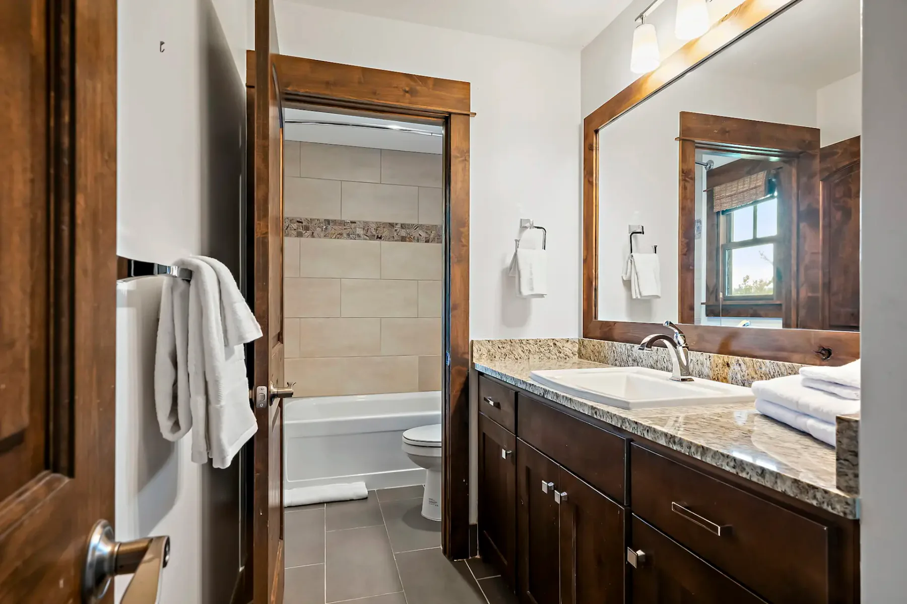 Baño moderno con encimera de granito, espejo grande, armarios de madera oscura y vistas a una zona de bañera revestida de azulejos. Las toallas cuelgan ordenadamente de los toalleros.