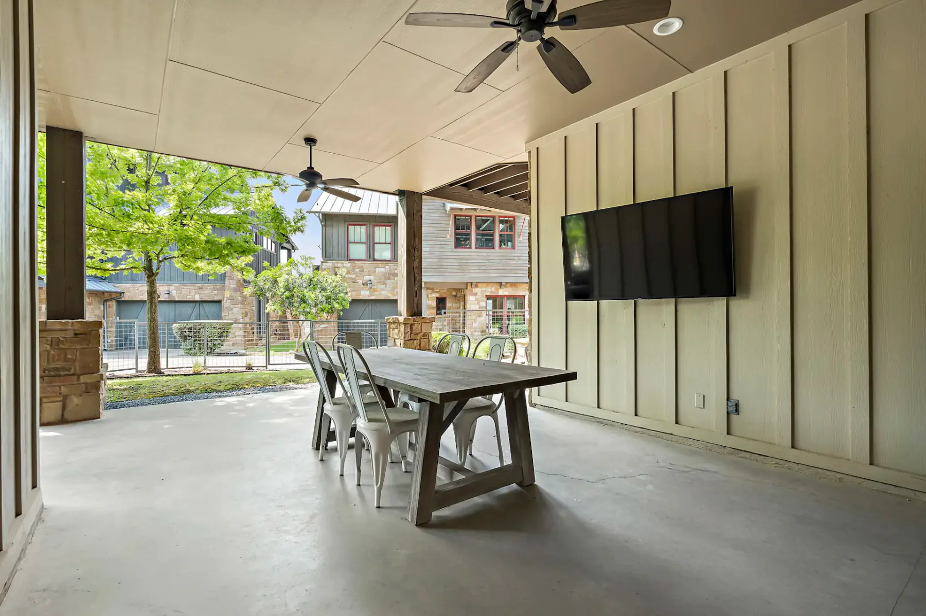 Patio cubierto con mesa de comedor de madera, sillas de metal, ventiladores de techo y televisor montado en la pared, con vistas a árboles y casas.