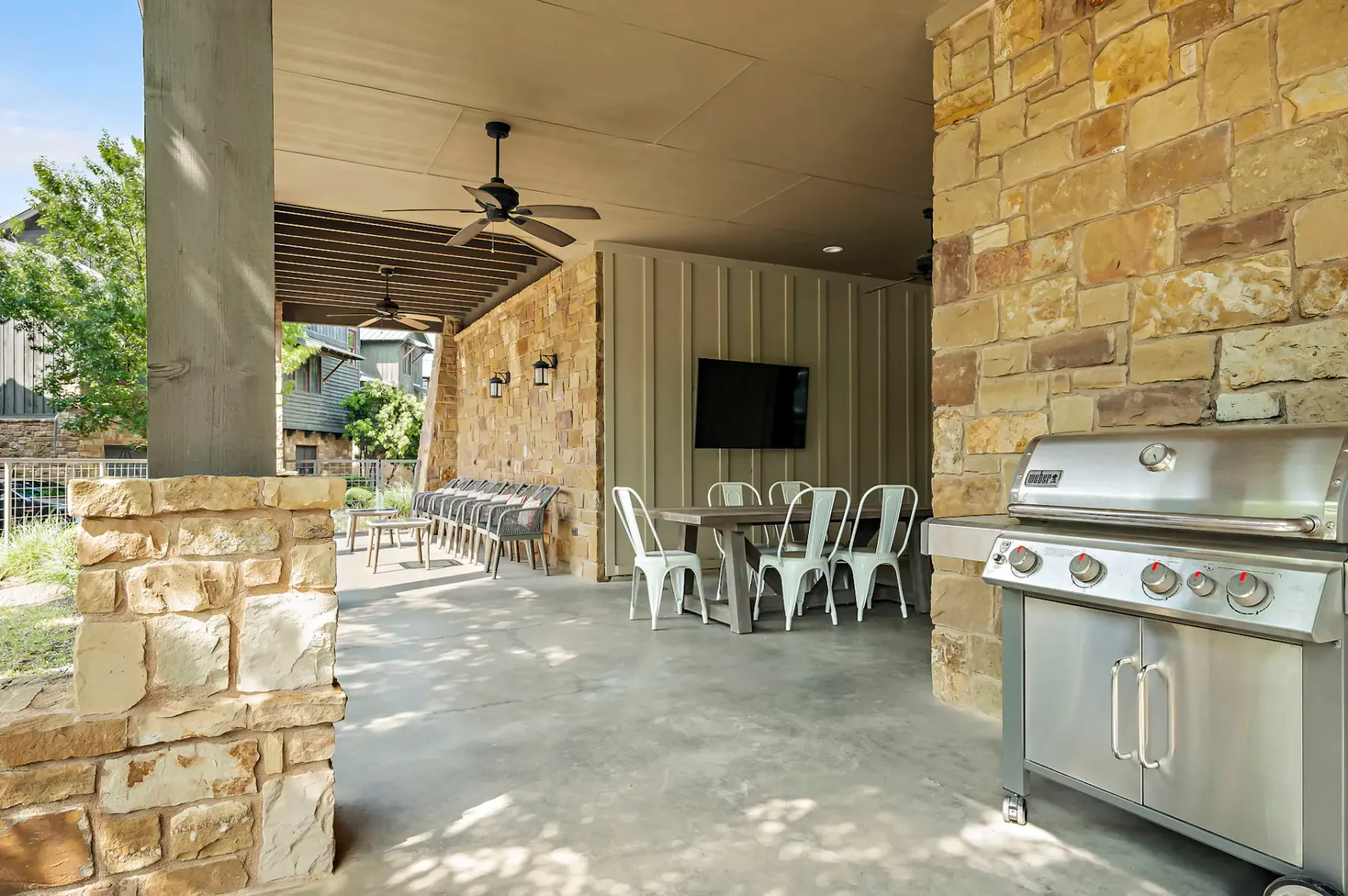 Patio exterior con paredes de piedra, parrillas y ventiladores de techo. Cuenta con una zona de comedor con sillas de metal y un televisor montado en la pared, bajo un techo que proporciona sombra.