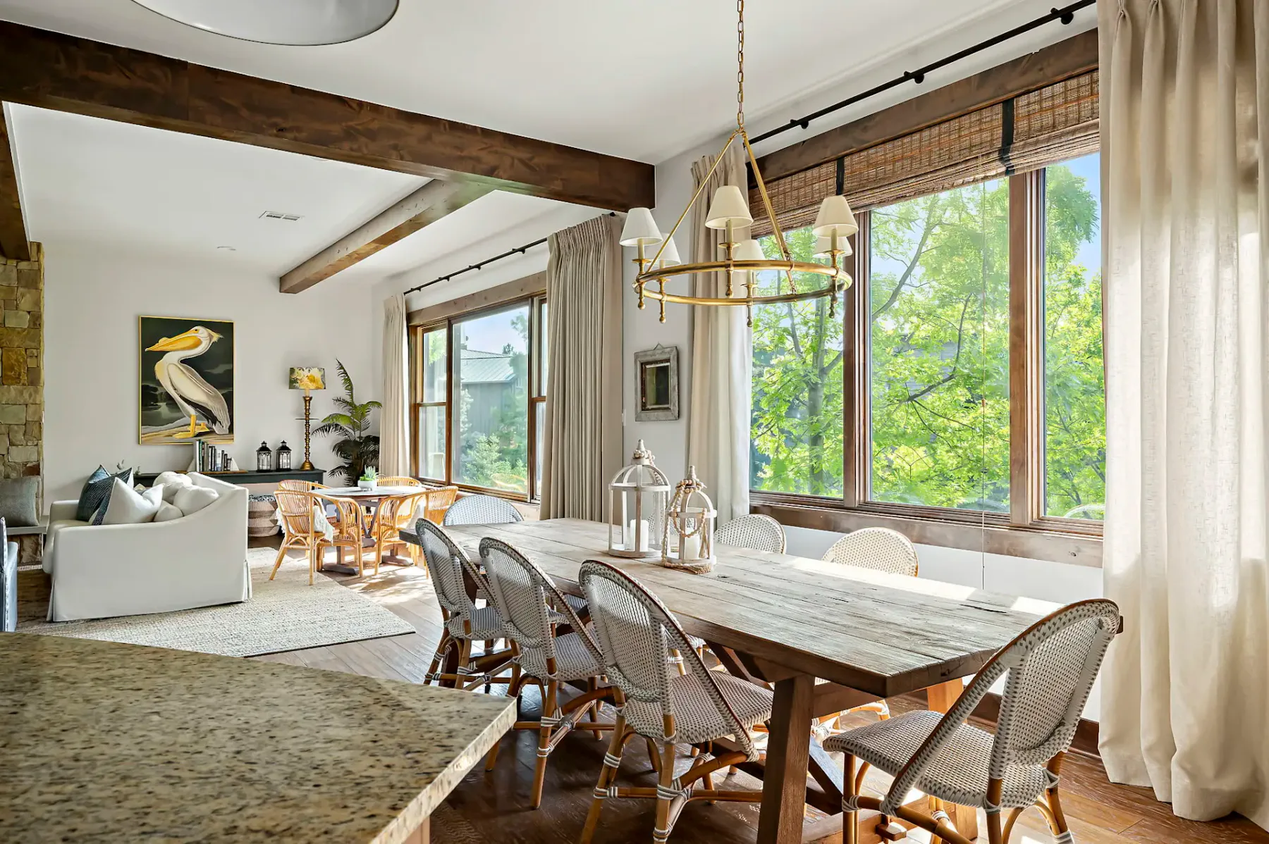 Bright dining room with a rustic wooden table, wicker chairs, large windows, beige curtains, and a cozy seating area in the background.