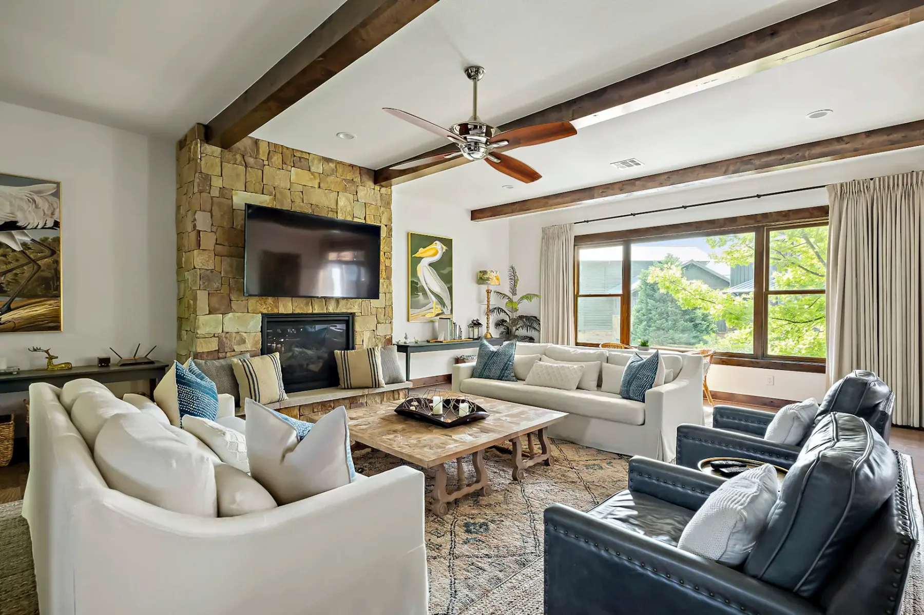 Acogedora sala de estar con chimenea de piedra, muebles modernos, ventilador de techo y una gran ventana con vistas a la exuberante vegetación exterior.